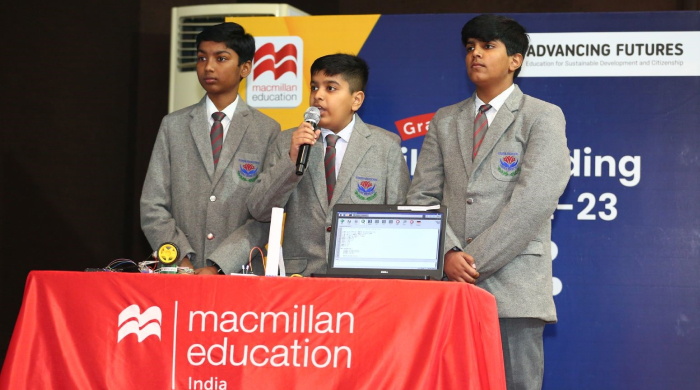 Three Indian boys standing in front of a table with a Macmillan Education tablecloth. They are all wearing school uniforms. One is talking into a microphone and others are presenting the project with a laptop.