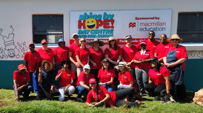 Group of people of various genders and ethnicities in red t-shirts smiling at the camera. They are standing in front of a banner for Shine by Hope! Day centre that is sponsored by Macmillan Education.