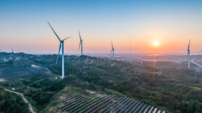 A landscape with four wind turbines and green hills with the sun setting in the background.