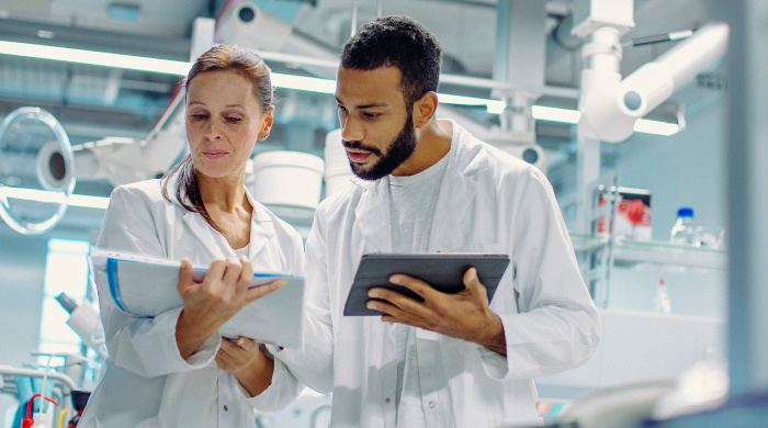 A man and woman working in a lab setting, both wearing lab coats and looking at information together on a tablet.