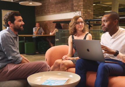 A Black man with a laptop open on his lap speaks to a white man and woman in a modern co-working space, sitting around a coffee table.