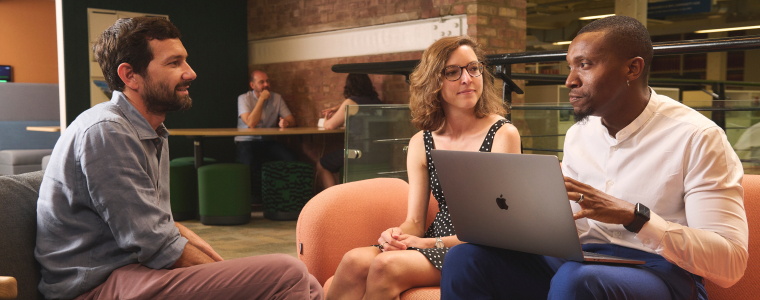 A Black man with a laptop open on his lap speaks to a white man and woman in a modern co-working space, sitting around a coffee table.