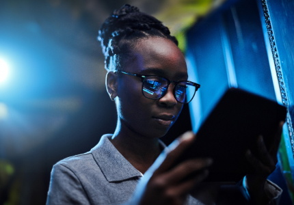A Black woman looking at a tablet with the screen reflecting in her glasses.